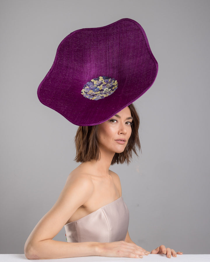The female model is posing, wearing
Ulma, the purple sinamay statement fascinator with an organically sculpted, flowing silhouette, featuring a central cluster of delicate yellow and violet flowers for a vibrant, artistic finish. Designed and made by Merve Bayindir Millinery.