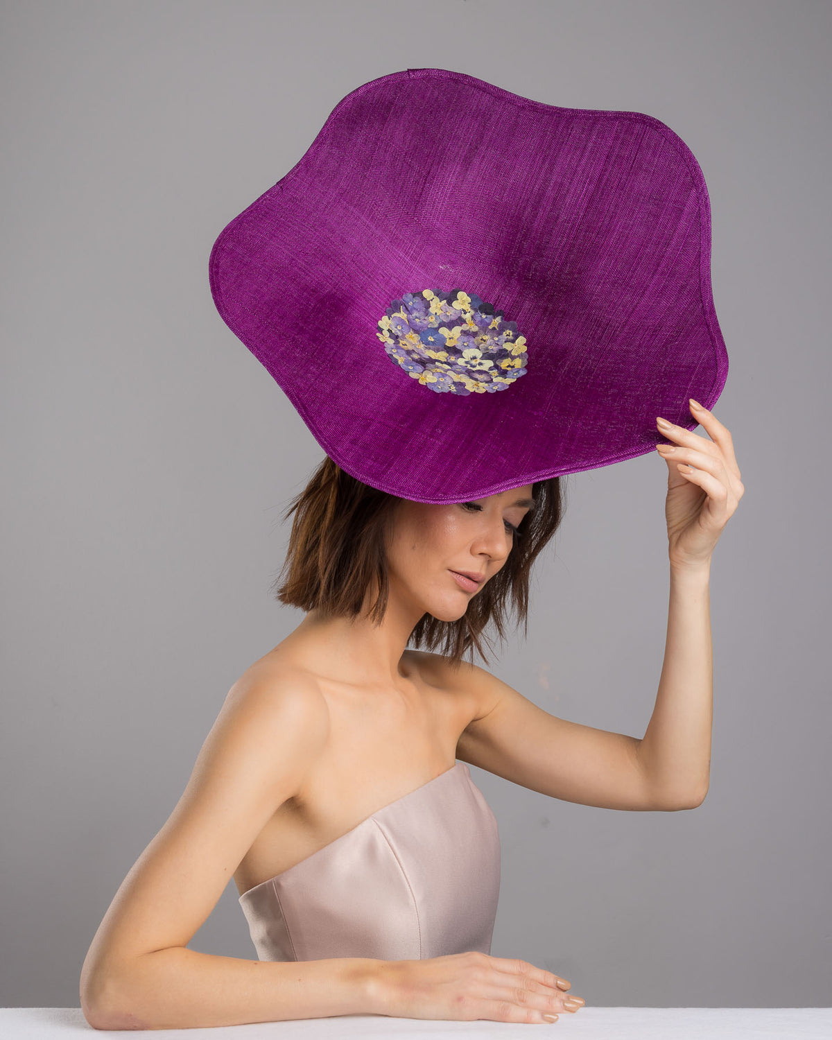 The female model is posing, wearing
Ulma, the purple sinamay statement fascinator with an organically sculpted, flowing silhouette, featuring a central cluster of delicate yellow and violet flowers for a vibrant, artistic finish. Designed and made by Merve Bayindir Millinery.

