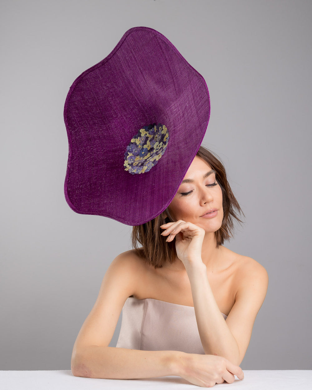 The female model is posing, wearing
Ulma, the purple sinamay statement fascinator with an organically sculpted, flowing silhouette, featuring a central cluster of delicate yellow and violet flowers for a vibrant, artistic finish. Designed and made by Merve Bayindir Millinery.