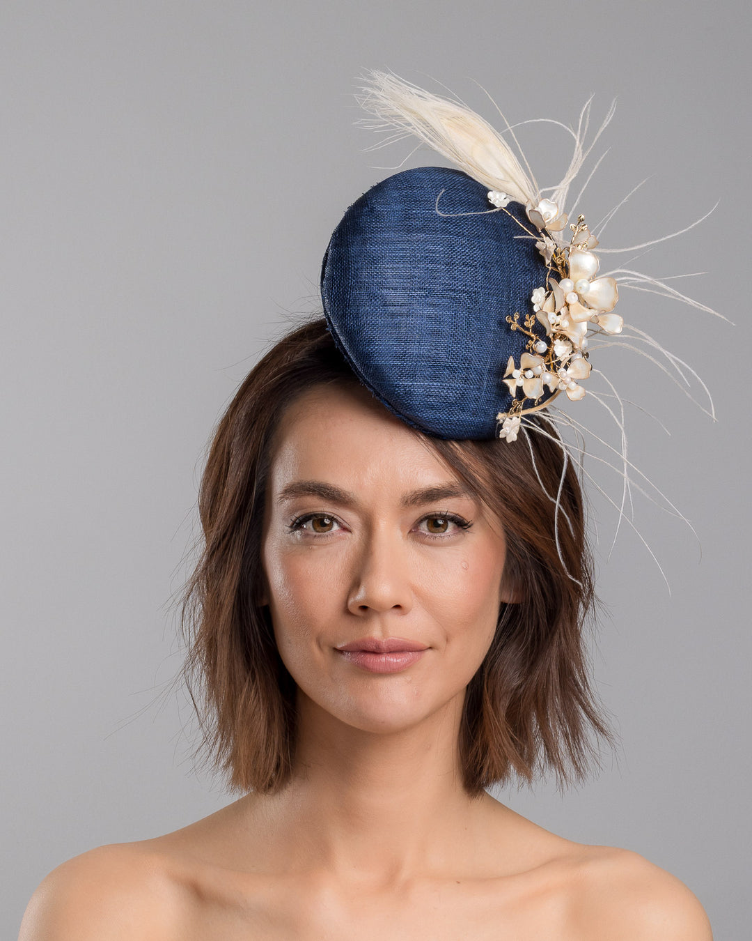 The model is showcasing the front side close up of the navy blue sinamay fascinator with a smooth rounded base, adorned with ivory floral embellishments, pearl accents, delicate gold branches, and soft white feather detailing for an elegant, refined finish. Designed and made by Merve Bayindir Millinery
