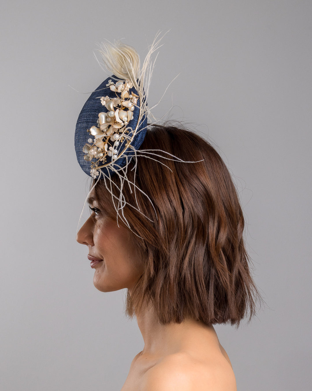 The model is showcasing the left side close up of the navy blue sinamay fascinator with a smooth rounded base, adorned with ivory floral embellishments, pearl accents, delicate gold branches, and soft white feather detailing for an elegant, refined finish. Designed and made by Merve Bayindir Millinery
