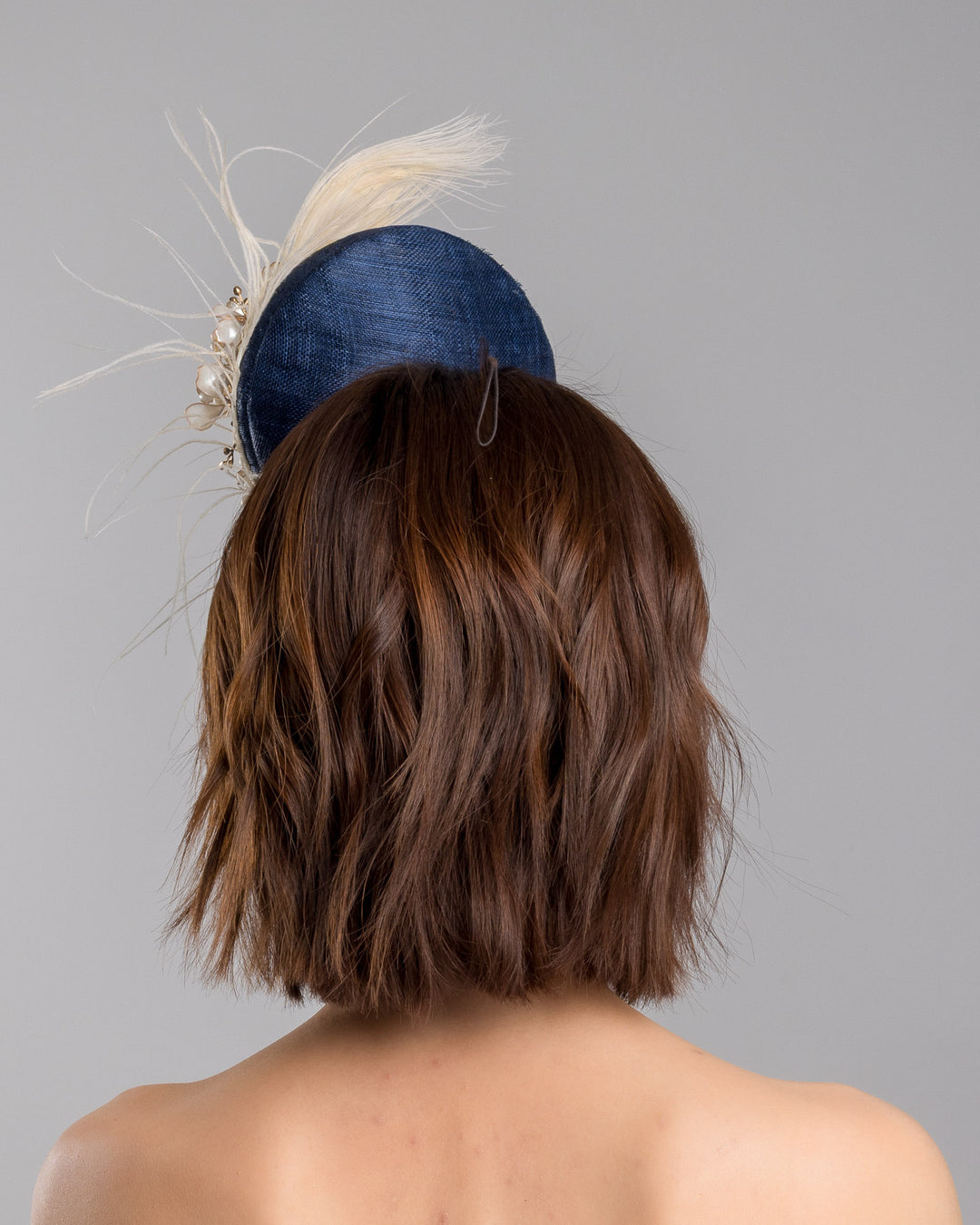 The model is showcasing the back side close up of the navy blue sinamay fascinator with a smooth rounded base, adorned with ivory floral embellishments, pearl accents, delicate gold branches, and soft white feather detailing for an elegant, refined finish. Designed and made by Merve Bayindir Millinery
