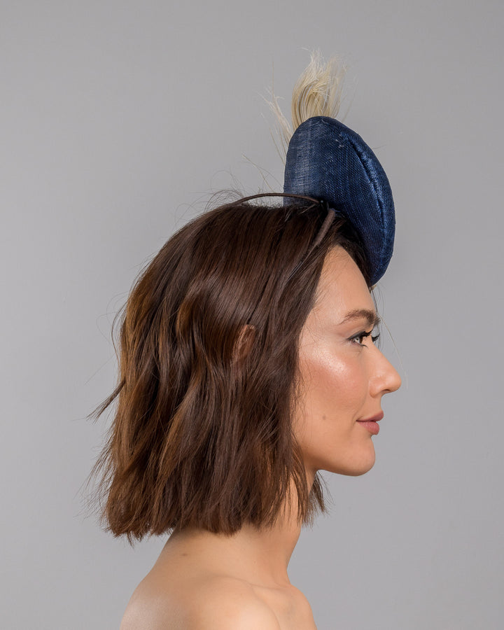 The model is showcasing the right side close up of the navy blue sinamay fascinator with a smooth rounded base, adorned with ivory floral embellishments, pearl accents, delicate gold branches, and soft white feather detailing for an elegant, refined finish. Designed and made by Merve Bayindir Millinery

