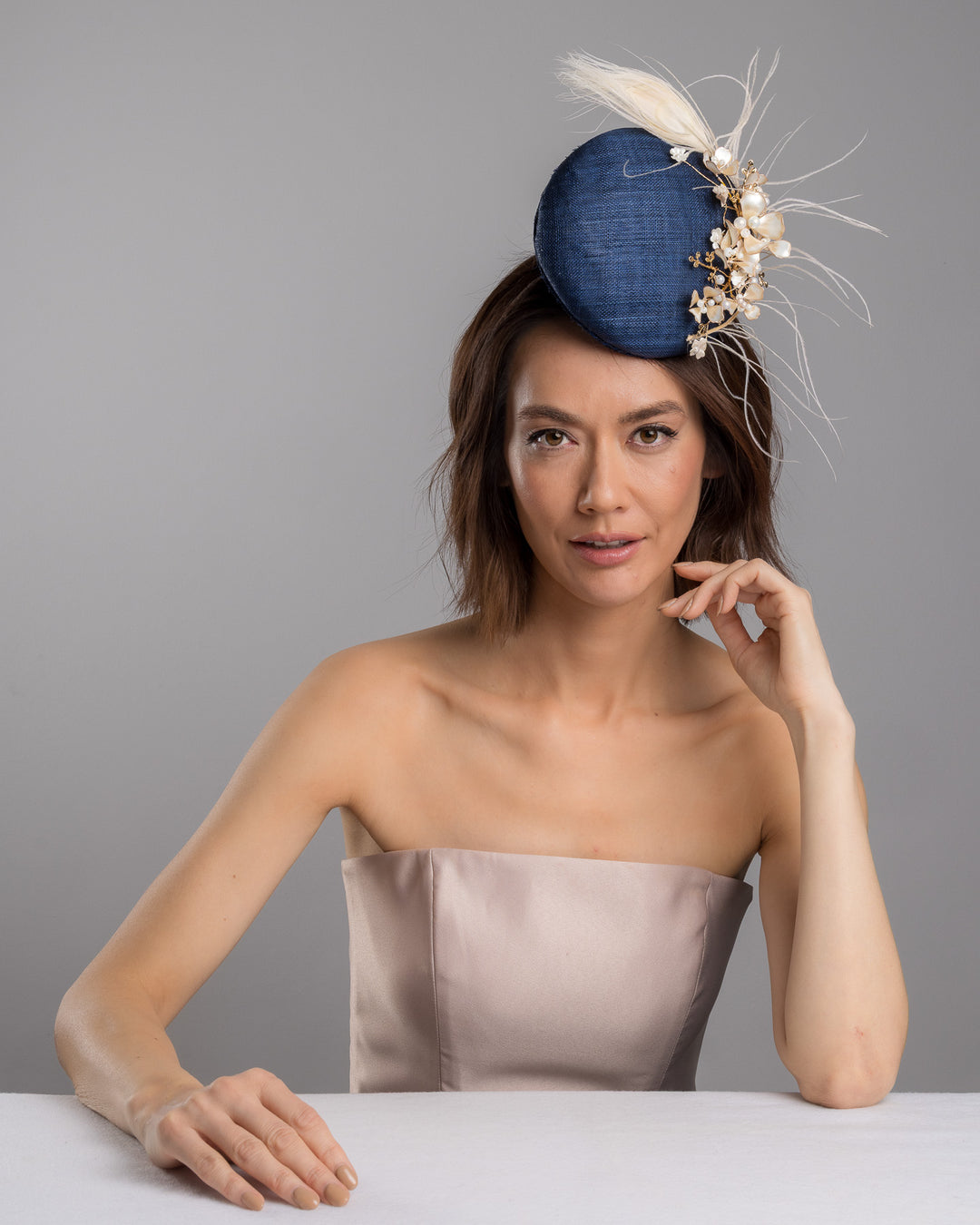 The female model is posing, wearing
Umber, the navy blue sinamay fascinator with a smooth rounded base, adorned with ivory floral embellishments, pearl accents, delicate gold branches, and soft white feather detailing for an elegant, refined finish. Designed and made by Merve Bayindir Millinery
