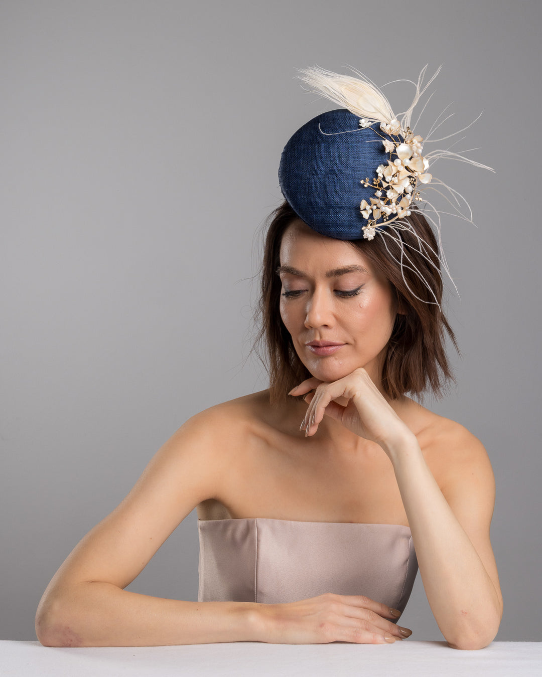 The female model is posing, wearing
Umber, the navy blue sinamay fascinator with a smooth rounded base, adorned with ivory floral embellishments, pearl accents, delicate gold branches, and soft white feather detailing for an elegant, refined finish. Designed and made by Merve Bayindir Millinery
