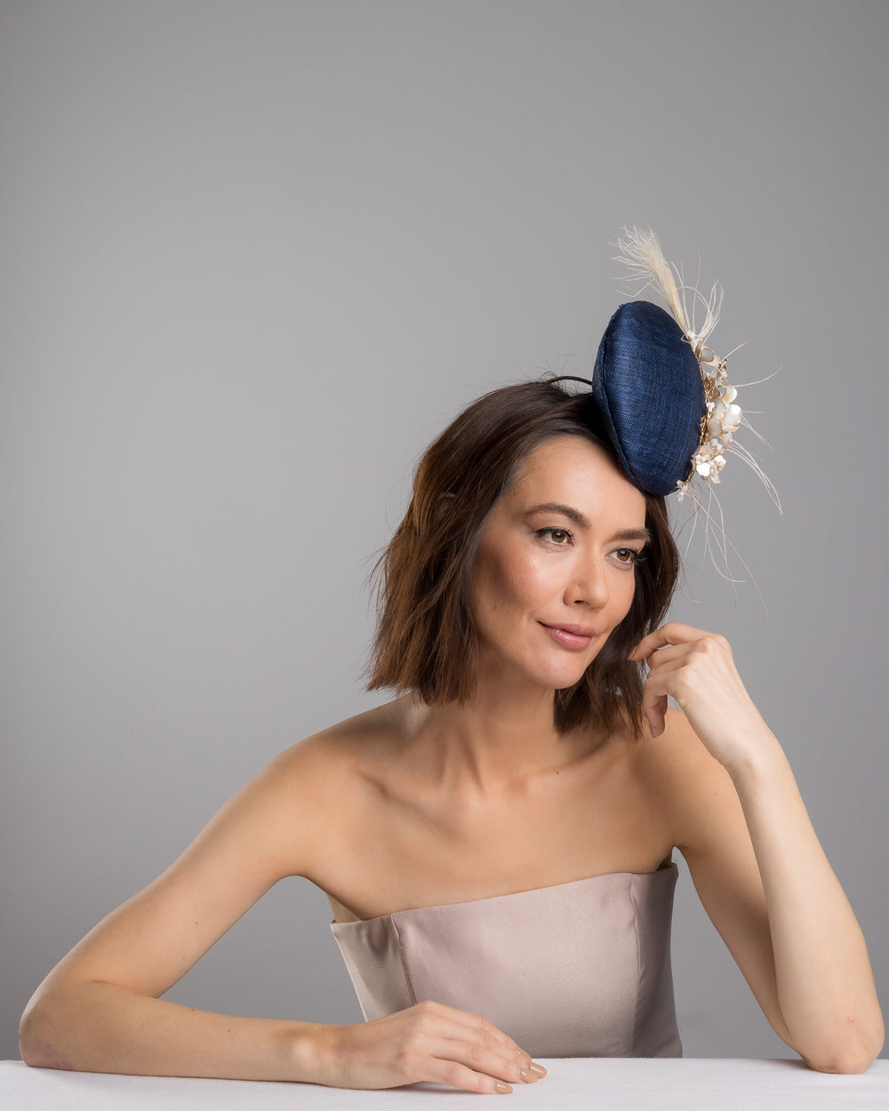 The female model is posing, wearing
Umber, the navy blue sinamay fascinator with a smooth rounded base, adorned with ivory floral embellishments, pearl accents, delicate gold branches, and soft white feather detailing for an elegant, refined finish. Designed and made by Merve Bayindir Millinery
