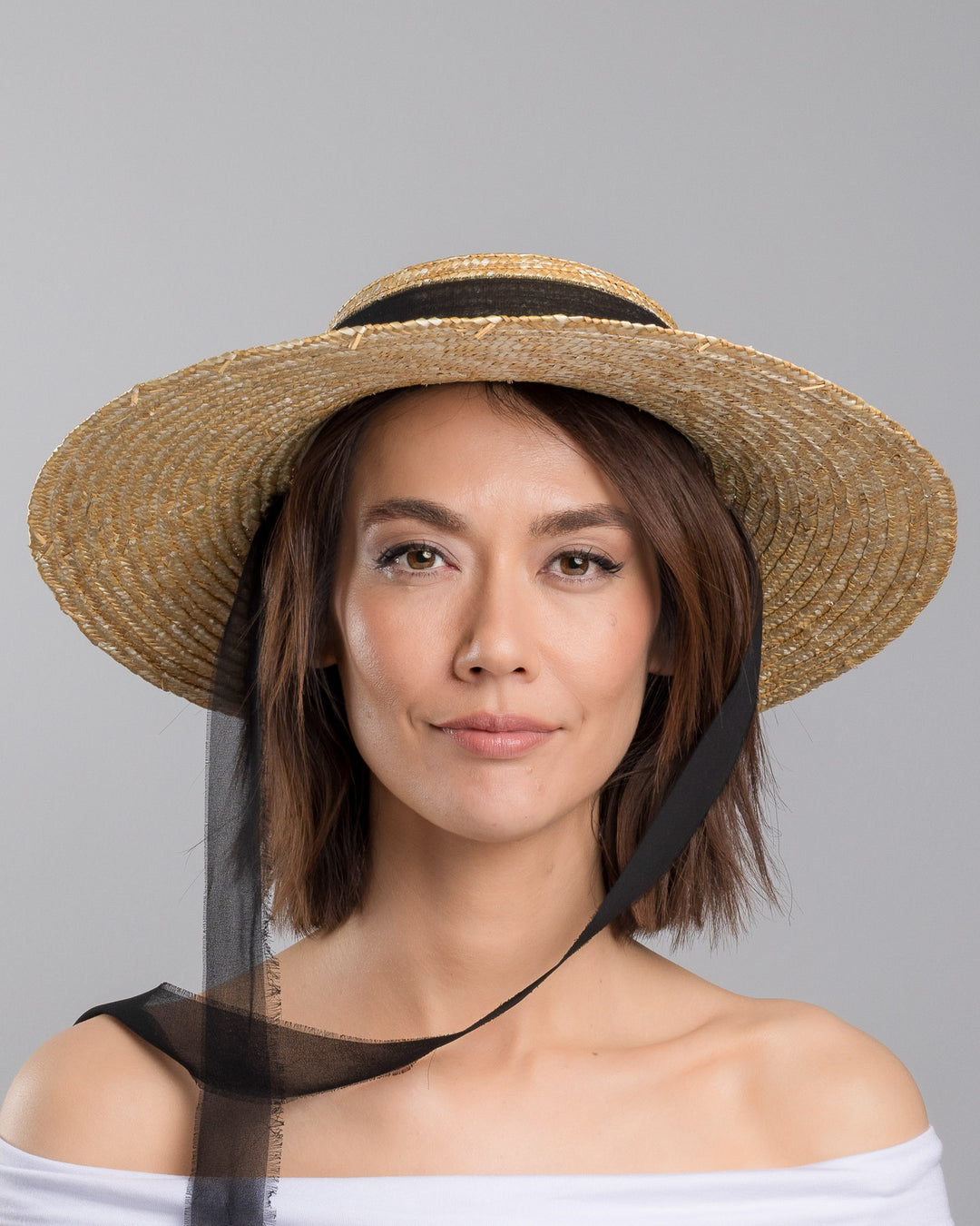The model is showcasing the front side close up of the natural woven straw boater hat with a wide brim, finished with a black ribbon band and long sheer black ties that extend from the back. Designed and made by Merve Bayindir millinery.