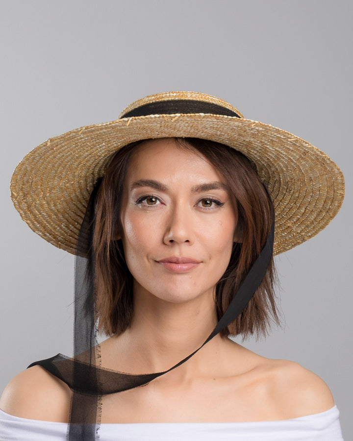 The model is showcasing the front side close up of the natural woven straw boater hat with a wide brim, finished with a black ribbon band and long sheer black ties that extend from the back. Designed and made by Merve Bayindir millinery.