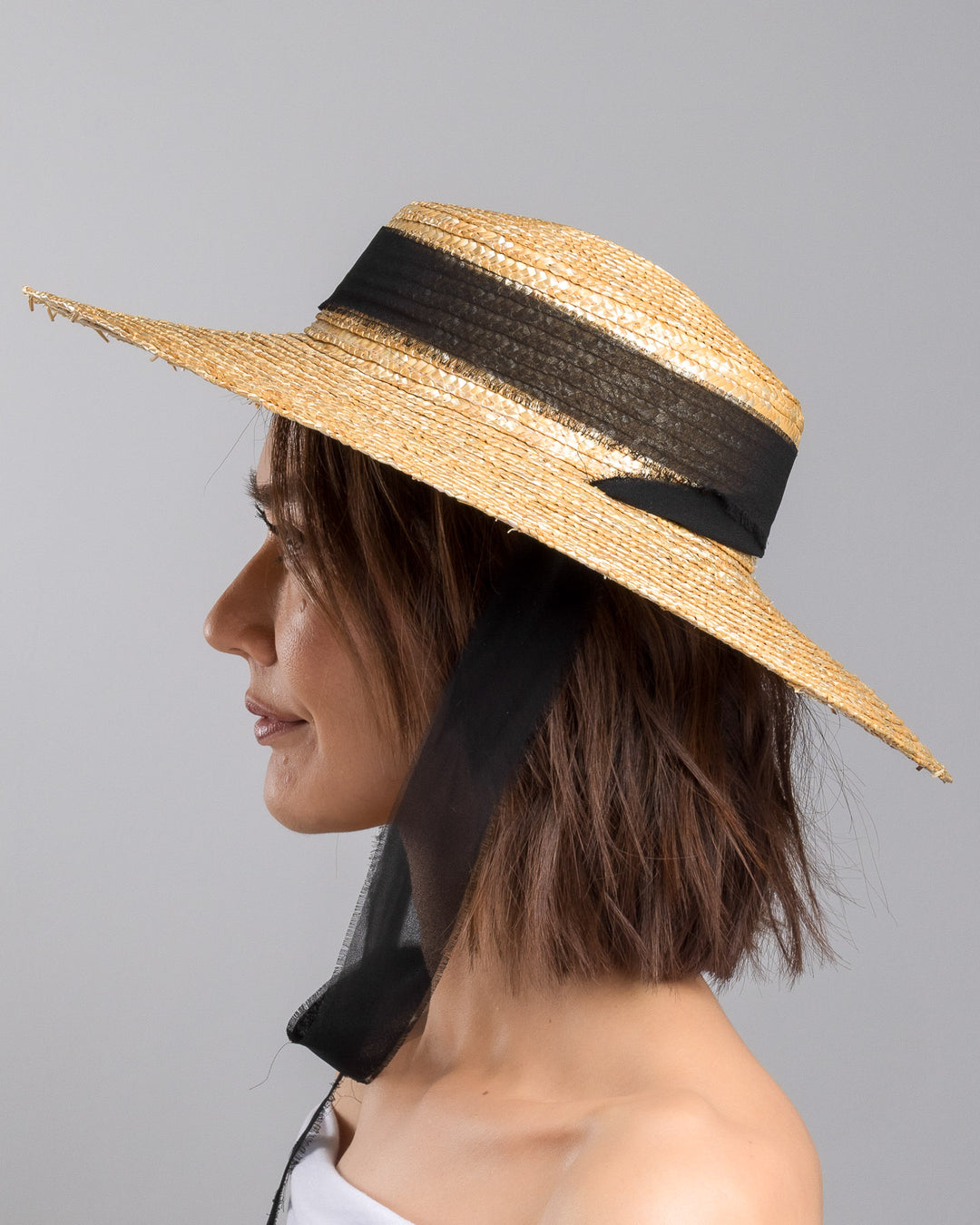 The model is showcasing the left side close up of the natural woven straw boater hat with a wide brim, finished with a black ribbon band and long sheer black ties that extend from the back. Designed and made by Merve Bayindir millinery.