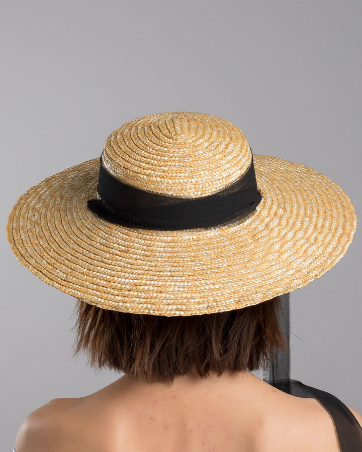 The model is showcasing the back side close up of the natural woven straw boater hat with a wide brim, finished with a black ribbon band and long sheer black ties that extend from the back. Designed and made by Merve Bayindir millinery.