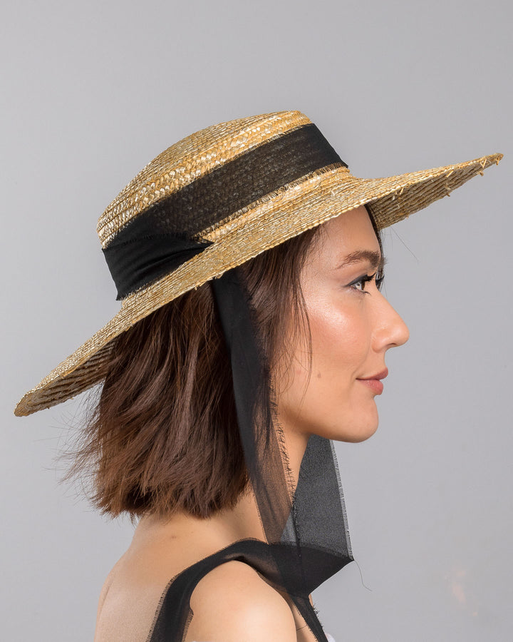 The model is showcasing the right side close up of the natural woven straw boater hat with a wide brim, finished with a black ribbon band and long sheer black ties that extend from the back. Designed and made by Merve Bayindir millinery.