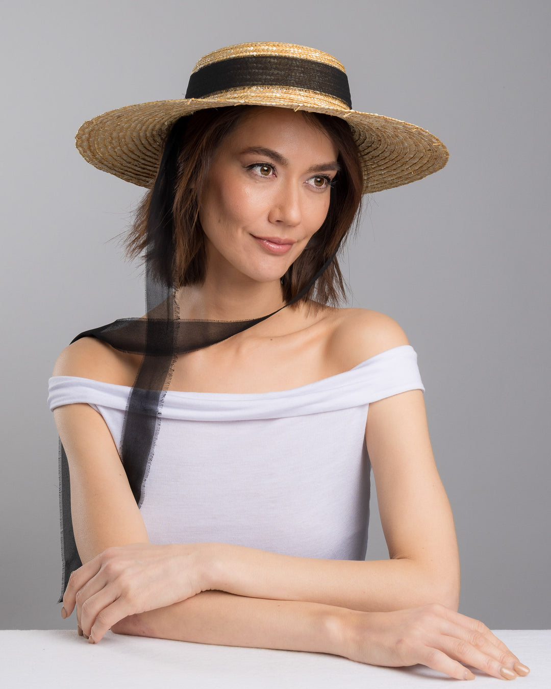 The female model is posing, wearing Vara, the natural woven straw boater hat with a wide brim, finished with a black ribbon band and long sheer black ties that extend from the back. Designed and made by Merve Bayindir millinery.