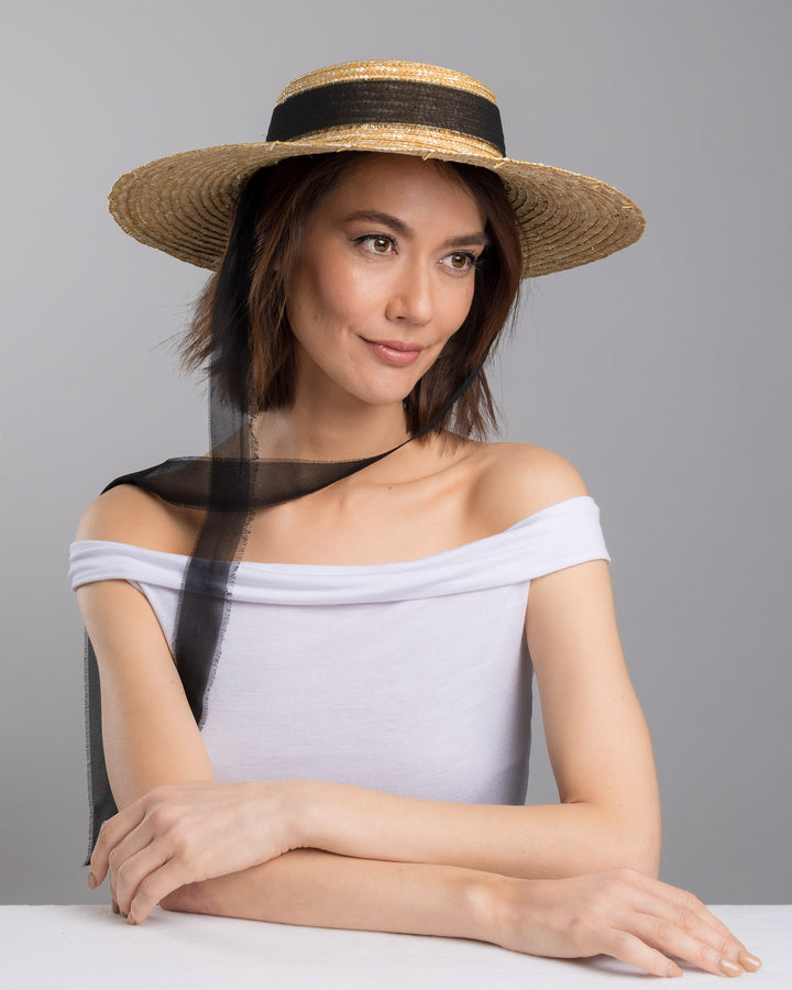 The female model is posing, wearing Vara, the natural woven straw boater hat with a wide brim, finished with a black ribbon band and long sheer black ties that extend from the back. Designed and made by Merve Bayindir millinery.