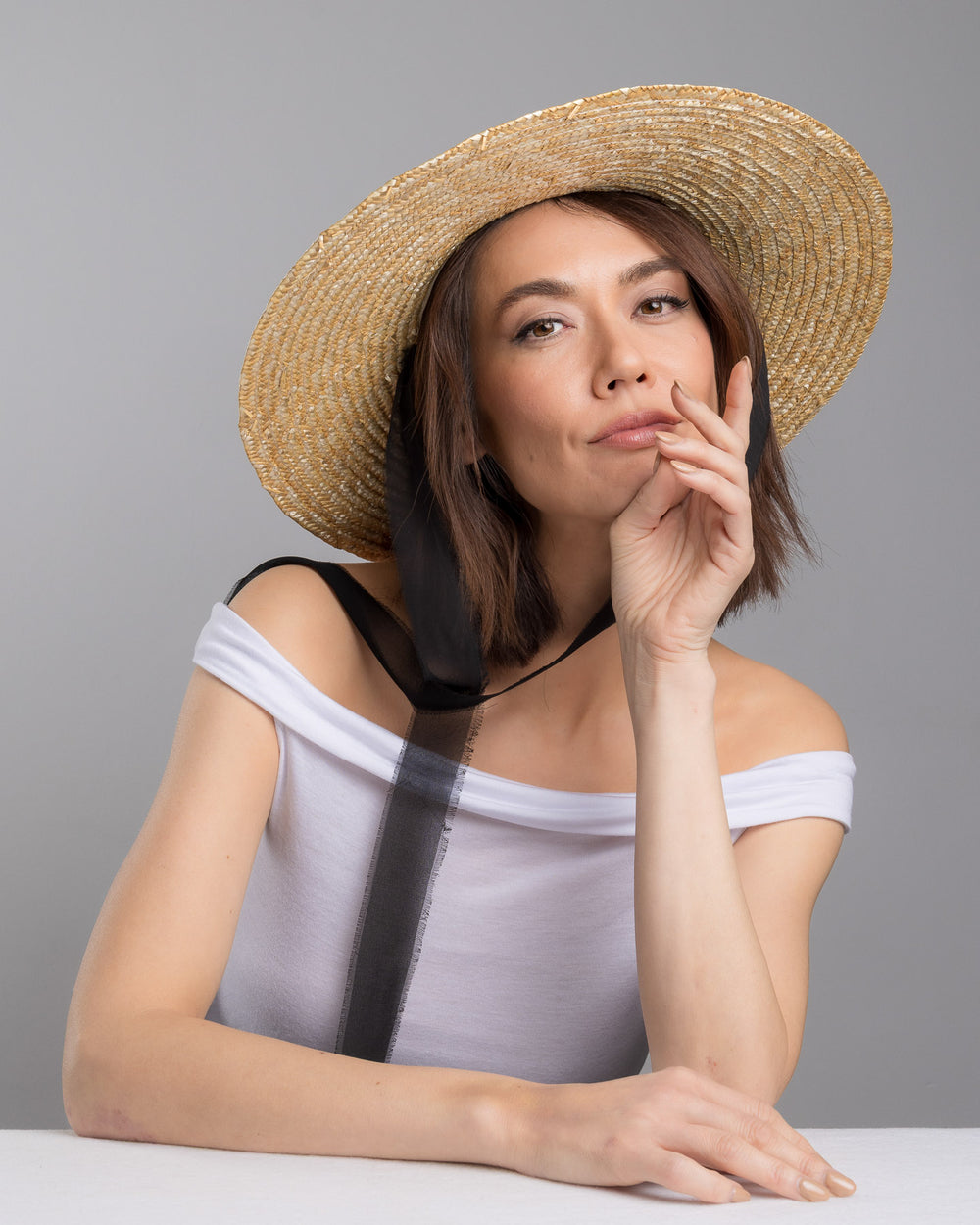 The female model is posing, wearing Vara, the natural woven straw boater hat with a wide brim, finished with a black ribbon band and long sheer black ties that extend from the back. Designed and made by Merve Bayindir millinery.