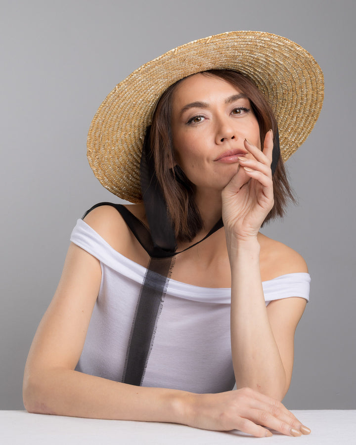 The female model is posing, wearing Vara, the natural woven straw boater hat with a wide brim, finished with a black ribbon band and long sheer black ties that extend from the back. Designed and made by Merve Bayindir millinery.