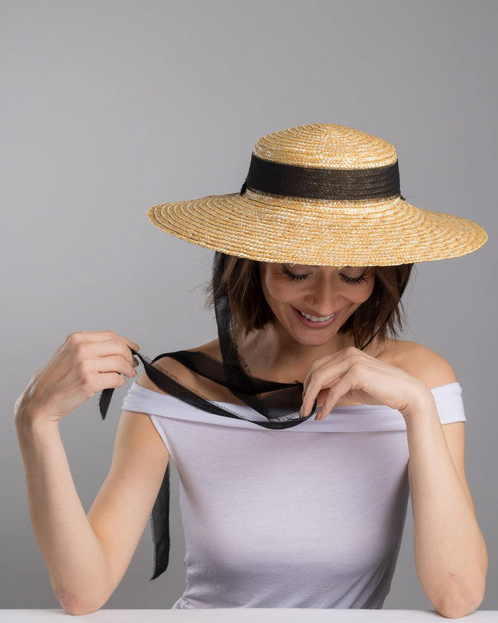 The female model is posing, wearing Vara, the natural woven straw boater hat with a wide brim, finished with a black ribbon band and long sheer black ties that extend from the back. Designed and made by Merve Bayindir millinery.