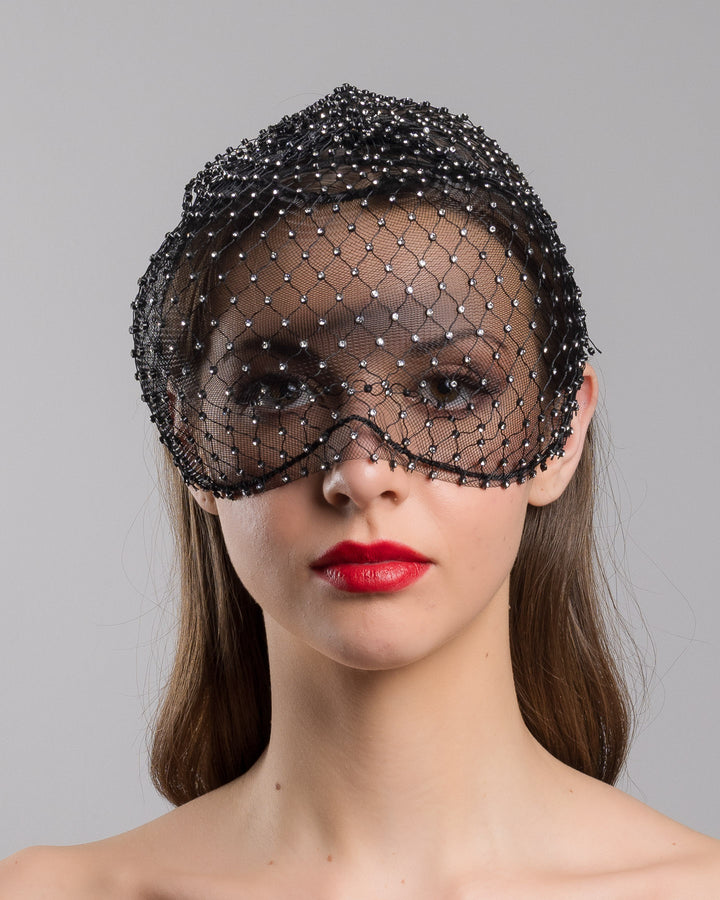 The model is showcasing the front side close up of the black birdcage veil mask crafted from fine netting, embellished with a grid of small silver crystal accents and trimmed with delicate beaded edging for a dramatic, couture-inspired finish. Designed and made by Merve Bayindir Millinery.