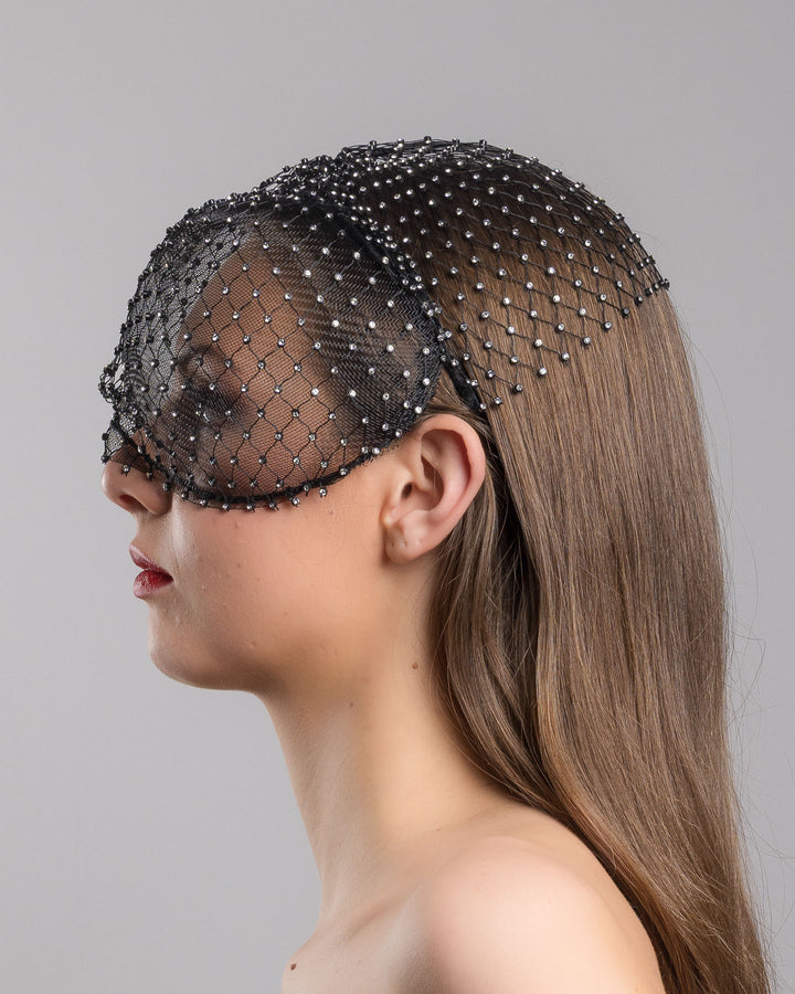 The model is showcasing the left side close up of the black birdcage veil mask crafted from fine netting, embellished with a grid of small silver crystal accents and trimmed with delicate beaded edging for a dramatic, couture-inspired finish. Designed and made by Merve Bayindir Millinery.