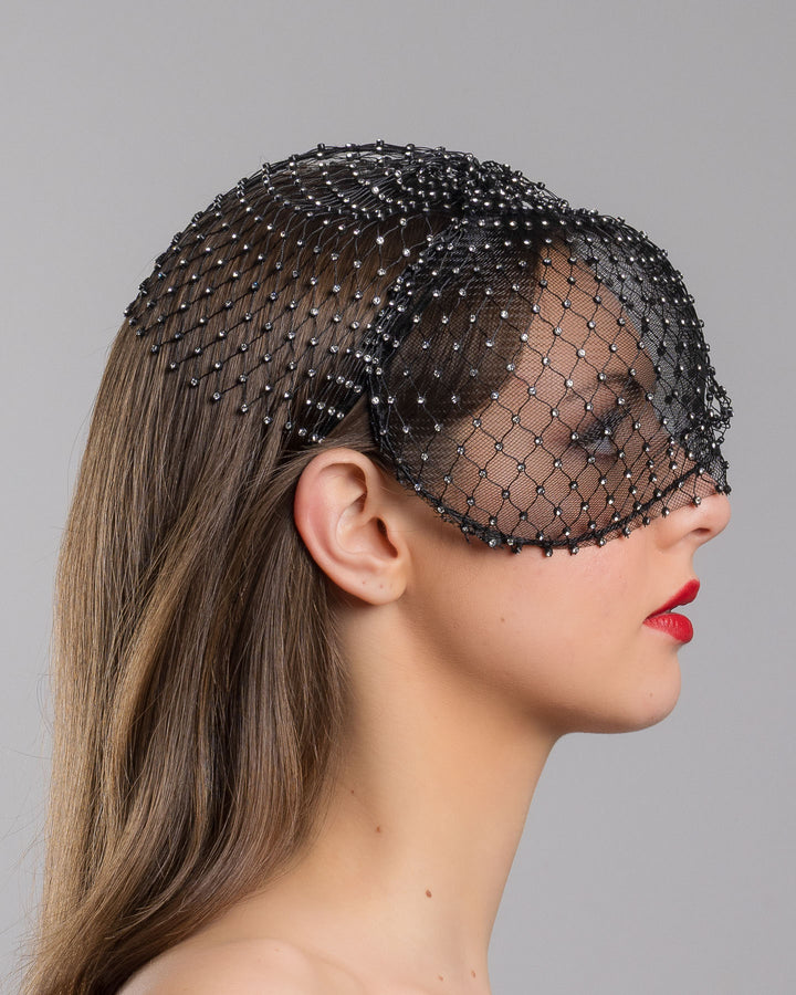 The model is showcasing the right side close up of the black birdcage veil mask crafted from fine netting, embellished with a grid of small silver crystal accents and trimmed with delicate beaded edging for a dramatic, couture-inspired finish. Designed and made by Merve Bayindir Millinery.
