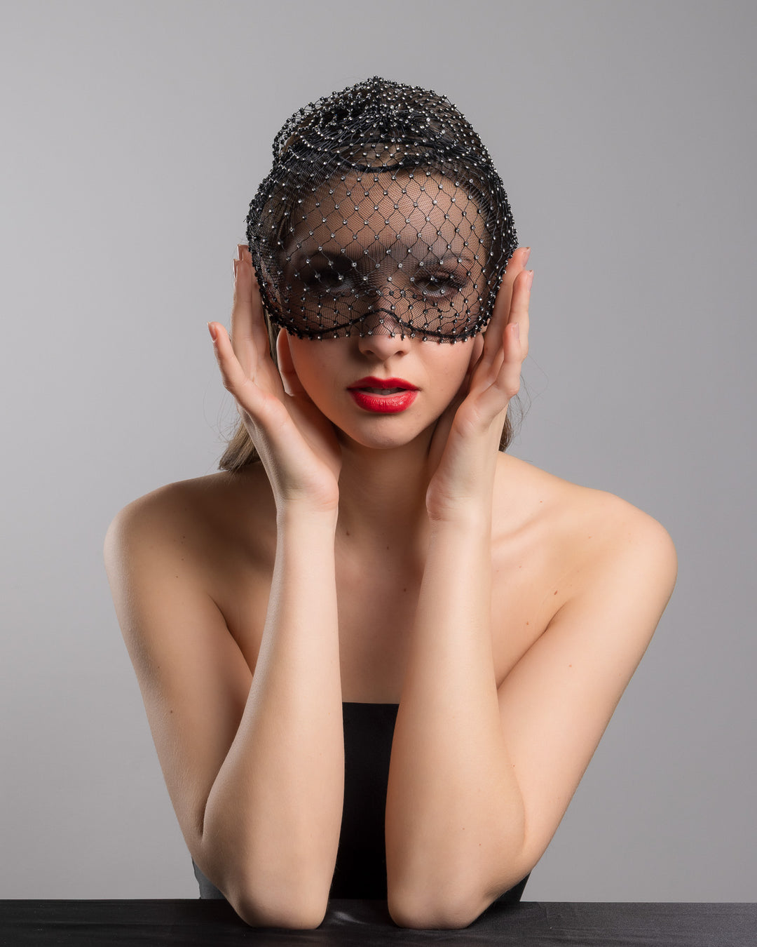The female model is posing, wearing 
Veir, the black birdcage veil mask crafted from fine netting, embellished with a grid of small silver crystal accents and trimmed with delicate beaded edging for a dramatic, couture-inspired finish. Designed and made by Merve Bayindir Millinery.