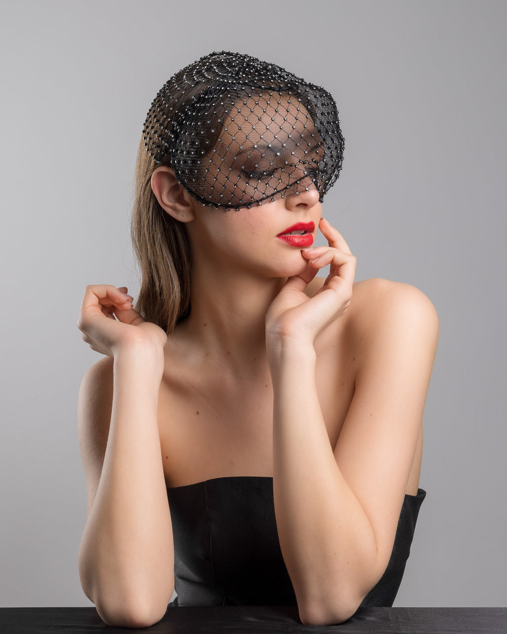 The female model is posing, wearing 
Veir, the black birdcage veil mask crafted from fine netting, embellished with a grid of small silver crystal accents and trimmed with delicate beaded edging for a dramatic, couture-inspired finish. Designed and made by Merve Bayindir Millinery.