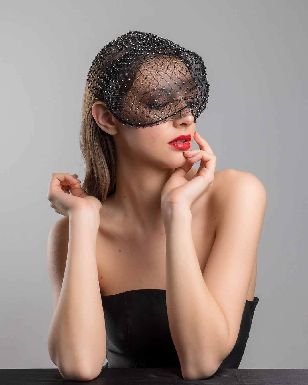 The female model is posing, wearing 
Veir, the black birdcage veil mask crafted from fine netting, embellished with a grid of small silver crystal accents and trimmed with delicate beaded edging for a dramatic, couture-inspired finish. Designed and made by Merve Bayindir Millinery.