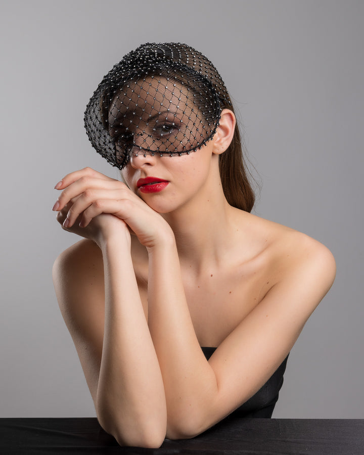 The female model is posing, wearing 
Veir, the black birdcage veil mask crafted from fine netting, embellished with a grid of small silver crystal accents and trimmed with delicate beaded edging for a dramatic, couture-inspired finish. Designed and made by Merve Bayindir Millinery.