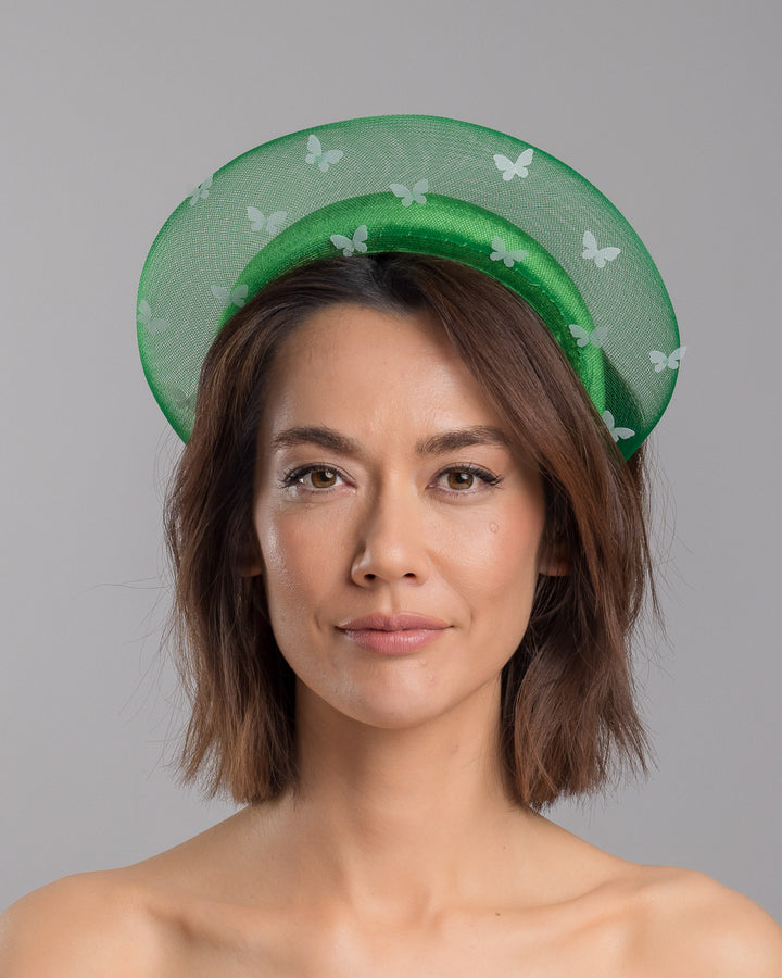 The model is showcasing the front side close up of the emerald green headband fascinator with a satin-covered base encircled by a sheer structured halo, decorated with small white butterfly appliqués for contrast and texture. Designed and made by Merve Bayindir Millinery.
