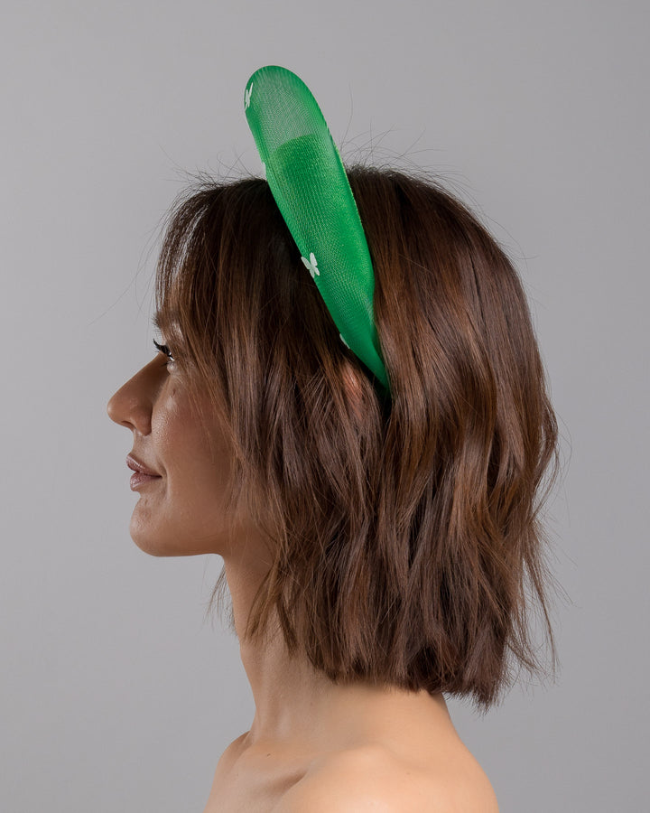 The model is showcasing the left side close up of the emerald green headband fascinator with a satin-covered base encircled by a sheer structured halo, decorated with small white butterfly appliqués for contrast and texture. Designed and made by Merve Bayindir Millinery.
