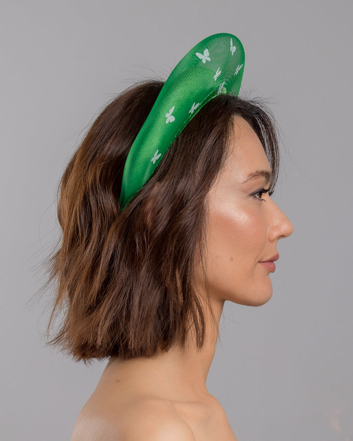 The model is showcasing the right side close up of the emerald green headband fascinator with a satin-covered base encircled by a sheer structured halo, decorated with small white butterfly appliqués for contrast and texture. Designed and made by Merve Bayindir Millinery.
