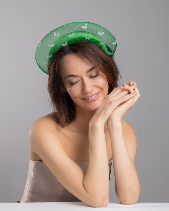 The female model is posing, wearing Verdel, the emerald green headband fascinator with a satin-covered base encircled by a sheer structured halo, decorated with small white butterfly appliqués for contrast and texture. Designed and made by Merve Bayindir Millinery.
