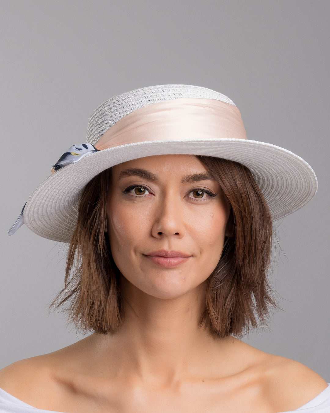 The model is showcasing the front side close up of the white woven straw boater hat with a wide brim, wrapped in a soft beige silk ribbon and finished with a tied scarf featuring illustrated panda motifs at the side. Designed and Made by Merve Bayindir millinery