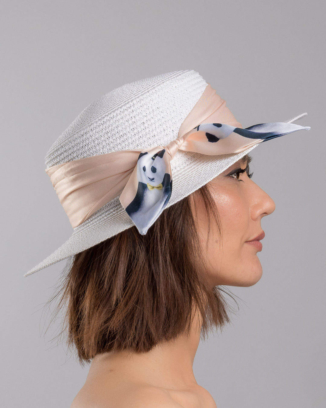 The model is showcasing the right side close up of the white woven straw boater hat with a wide brim, wrapped in a soft beige silk ribbon and finished with a tied scarf featuring illustrated panda motifs at the side. Designed and Made by Merve Bayindir millinery