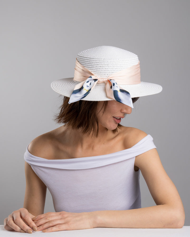 The female model is posing, wearing Vianne, the white woven straw boater hat with a wide brim, wrapped in a soft beige silk ribbon and finished with a tied scarf featuring illustrated panda motifs at the side. Designed and Made by Merve Bayindir millinery