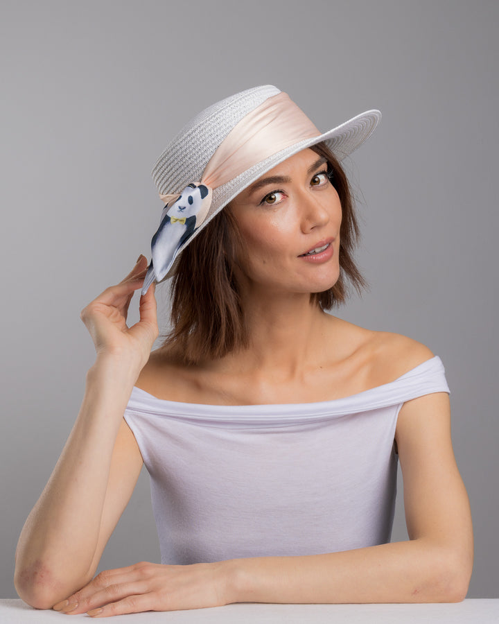 The female model is posing, wearing Vianne, the white woven straw boater hat with a wide brim, wrapped in a soft beige silk ribbon and finished with a tied scarf featuring illustrated panda motifs at the side. Designed and Made by Merve Bayindir millinery