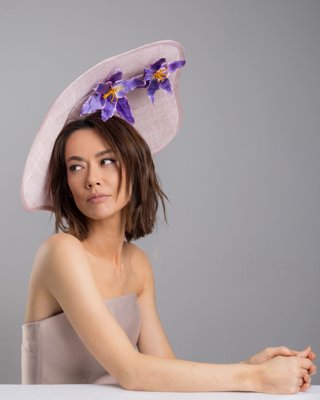 The female model is posing, wearing
Virel, the lilac sinamay wide-brim fascinator with a softly curved sculptural silhouette, featuring striking purple floral accents nestled beneath the brim for a bold yet refined finish. Designed and made by Merve Bayindir Millinery.
