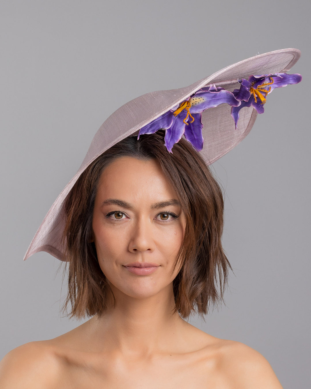 The model is showcasing the front side close up of the lilac sinamay wide-brim fascinator with a softly curved sculptural silhouette, featuring striking purple floral accents nestled beneath the brim for a bold yet refined finish. Designed and made by Merve Bayindir Millinery.
