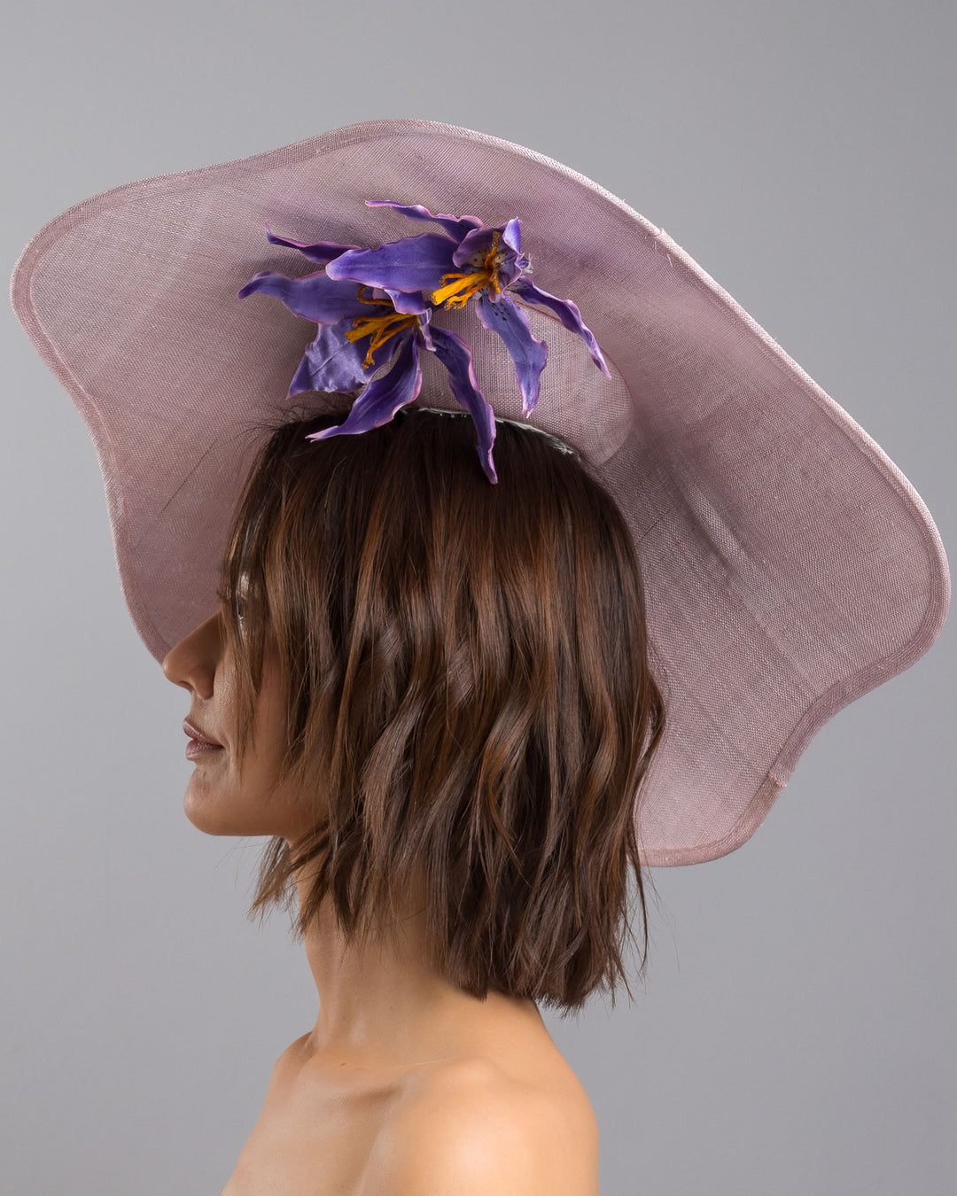 The model is showcasing the left side close up of the lilac sinamay wide-brim fascinator with a softly curved sculptural silhouette, featuring striking purple floral accents nestled beneath the brim for a bold yet refined finish. Designed and made by Merve Bayindir Millinery.

