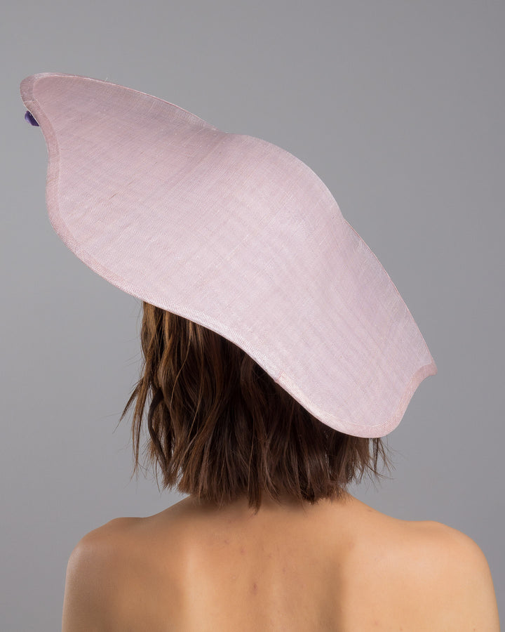 The model is showcasing the back side close up of the lilac sinamay wide-brim fascinator with a softly curved sculptural silhouette, featuring striking purple floral accents nestled beneath the brim for a bold yet refined finish. Designed and made by Merve Bayindir Millinery.
