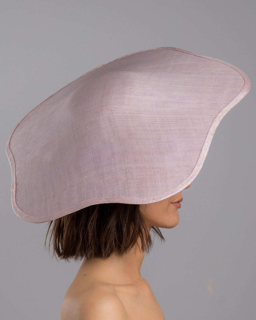 The model is showcasing the right side close up of the lilac sinamay wide-brim fascinator with a softly curved sculptural silhouette, featuring striking purple floral accents nestled beneath the brim for a bold yet refined finish. Designed and made by Merve Bayindir Millinery.
