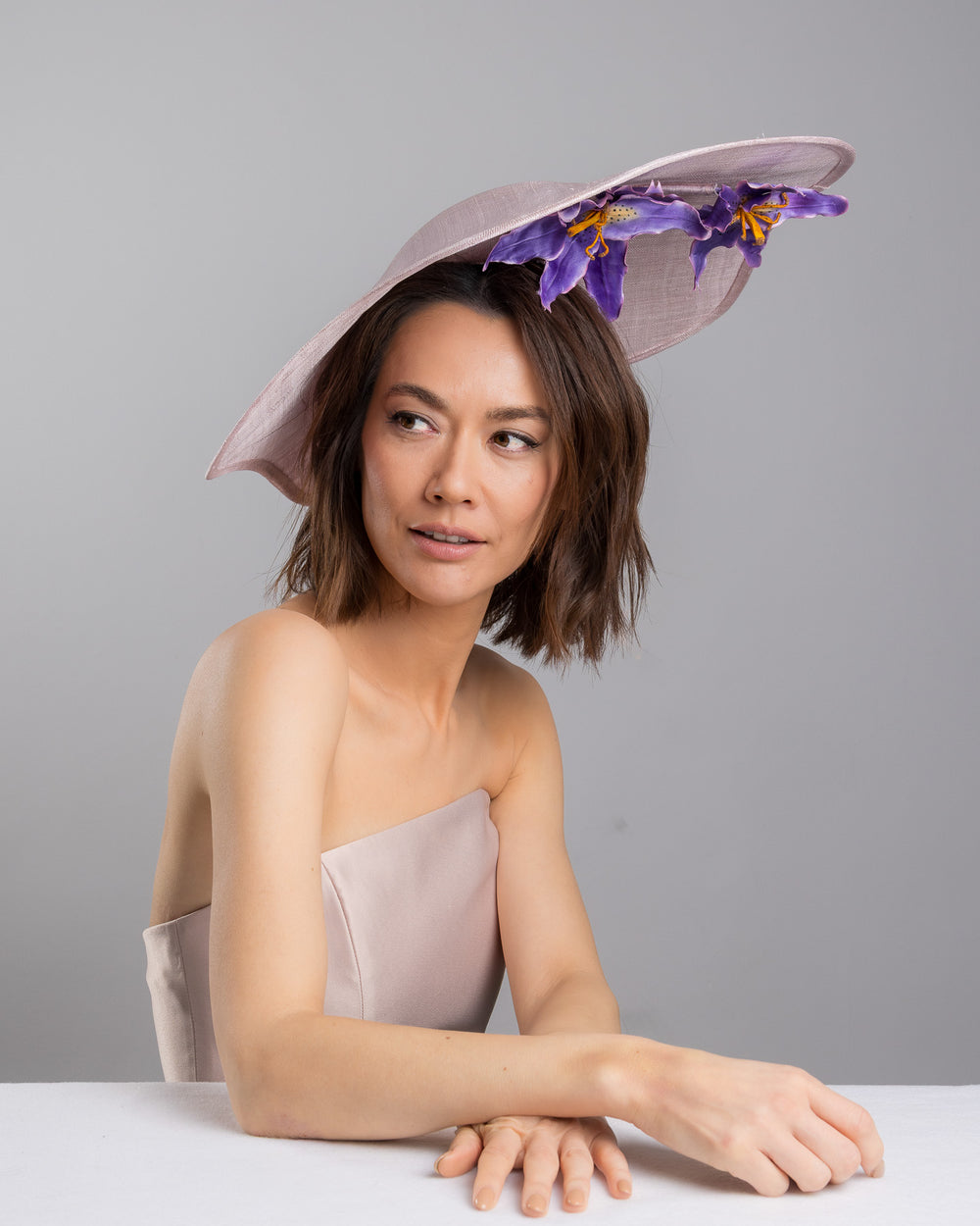 The female model is posing, wearing
Virel, the lilac sinamay wide-brim fascinator with a softly curved sculptural silhouette, featuring striking purple floral accents nestled beneath the brim for a bold yet refined finish. Designed and made by Merve Bayindir Millinery.
