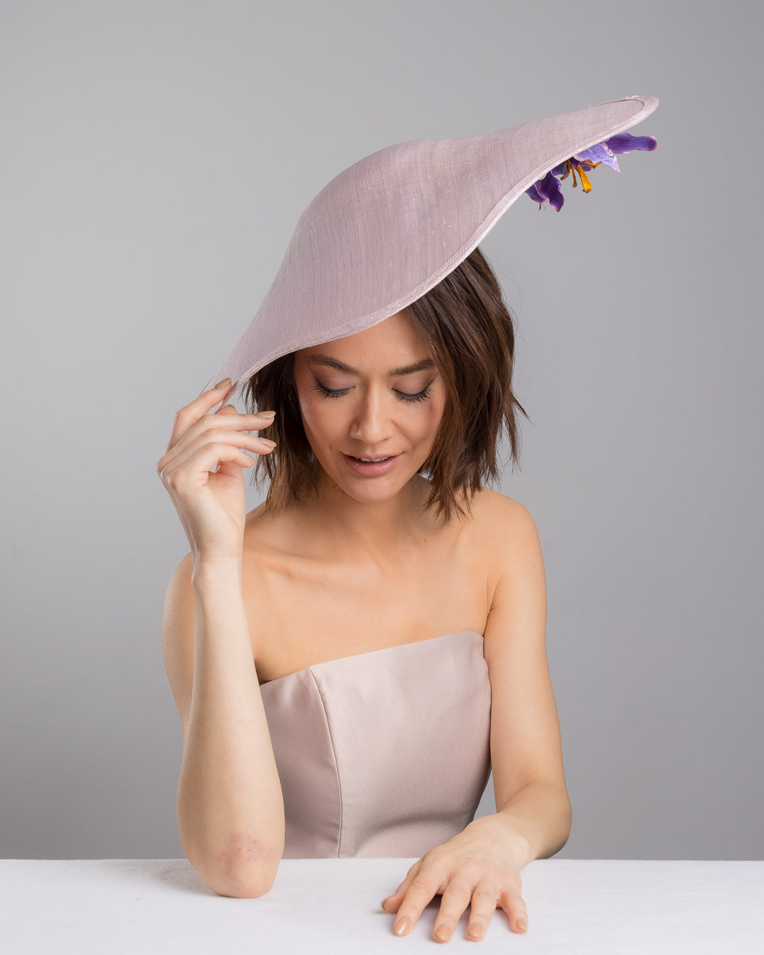 The female model is posing, wearing
Virel, the lilac sinamay wide-brim fascinator with a softly curved sculptural silhouette, featuring striking purple floral accents nestled beneath the brim for a bold yet refined finish. Designed and made by Merve Bayindir Millinery.
