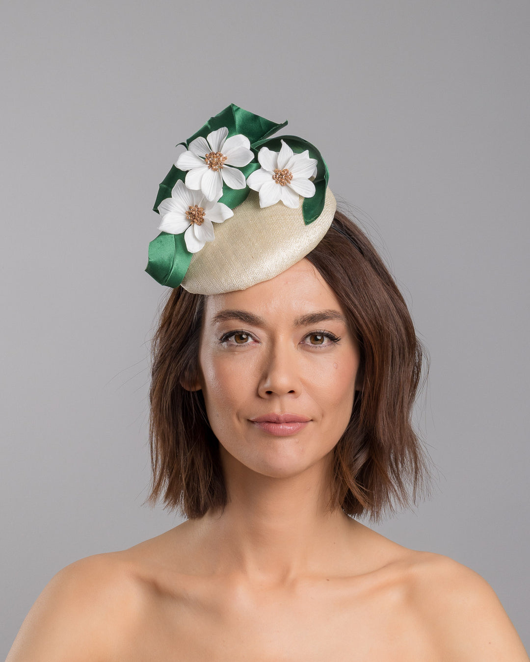 Model is showcasing the front side close up of Val, a button shaped, ivory sinamay fascinator worn to the front right side of the head. The fascinator is decorated with delicate white silk flowers and green sateen leaf swirls. Designed and made by Merve Bayindir millinery.