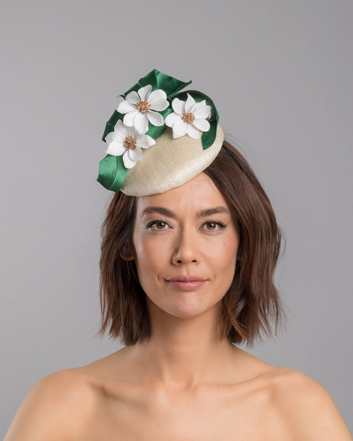 Model is showcasing the front side close up of Val, a button shaped, ivory sinamay fascinator worn to the front right side of the head. The fascinator is decorated with delicate white silk flowers and green sateen leaf swirls. Designed and made by Merve Bayindir millinery.