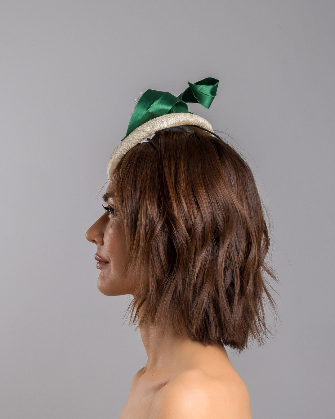 Model is showcasing the left side close up of Val, a button shaped, ivory sinamay fascinator worn to the front right side of the head. The fascinator is decorated with delicate white silk flowers and green sateen leaf swirls. Designed and made by Merve Bayindir millinery.