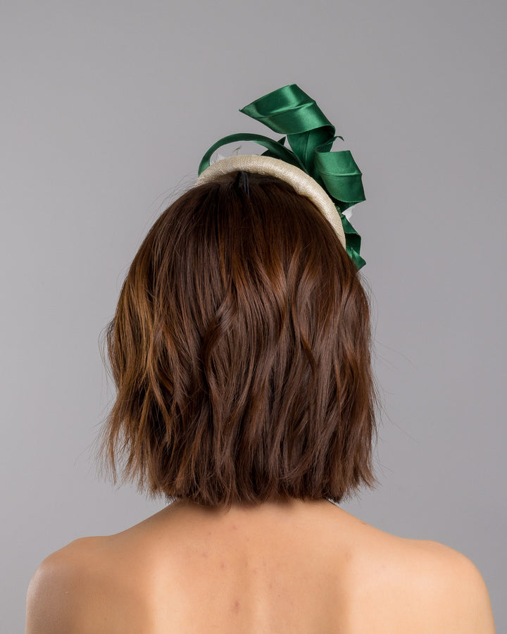 Model is showcasing the back side close up of Val, a button shaped, ivory sinamay fascinator worn to the front right side of the head. The fascinator is decorated with delicate white silk flowers and green sateen leaf swirls. Designed and made by Merve Bayindir millinery.