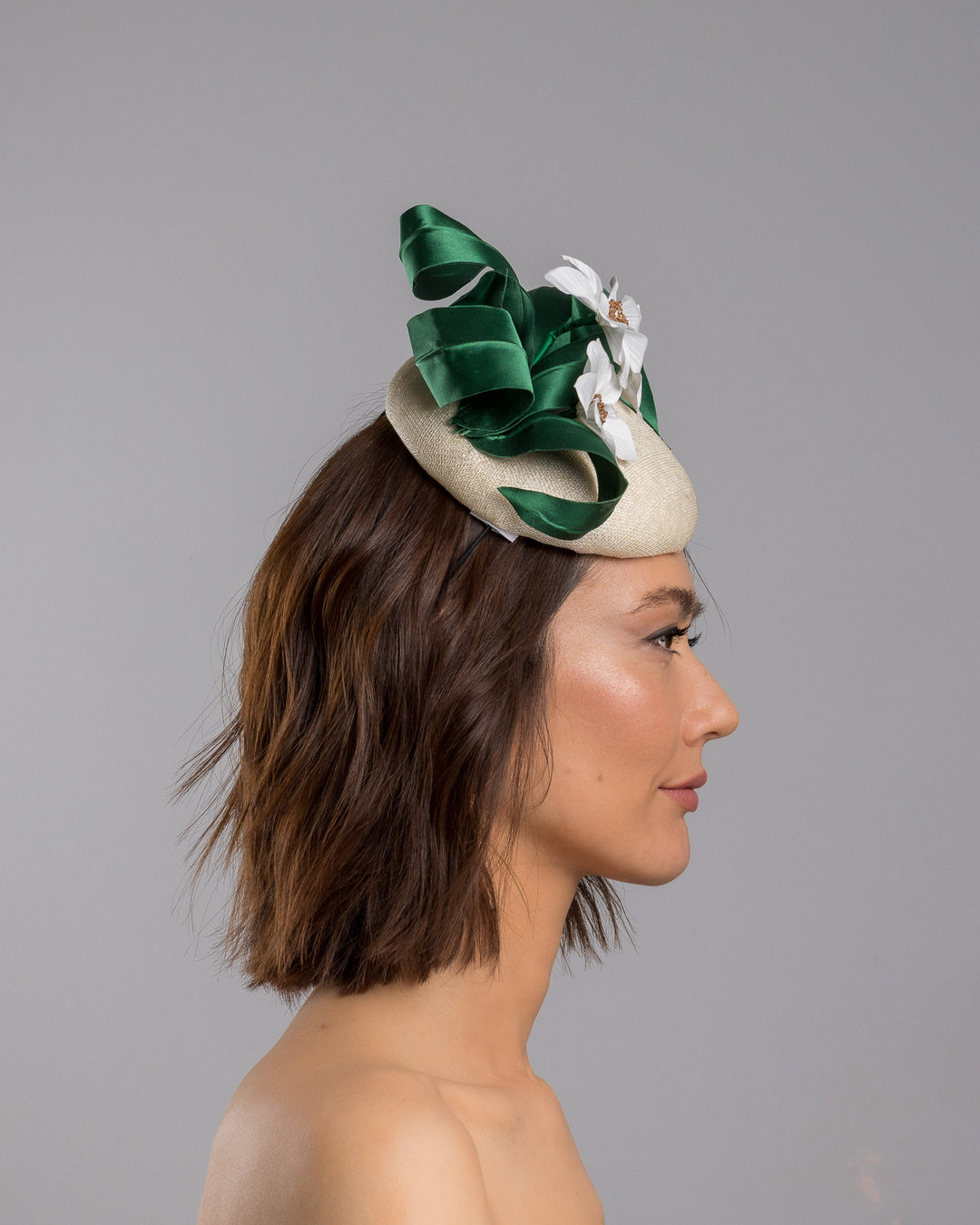 Model is showcasing the right side close up of Val, a button shaped, ivory sinamay fascinator worn to the front right side of the head. The fascinator is decorated with delicate white silk flowers and green sateen leaf swirls. Designed and made by Merve Bayindir millinery.