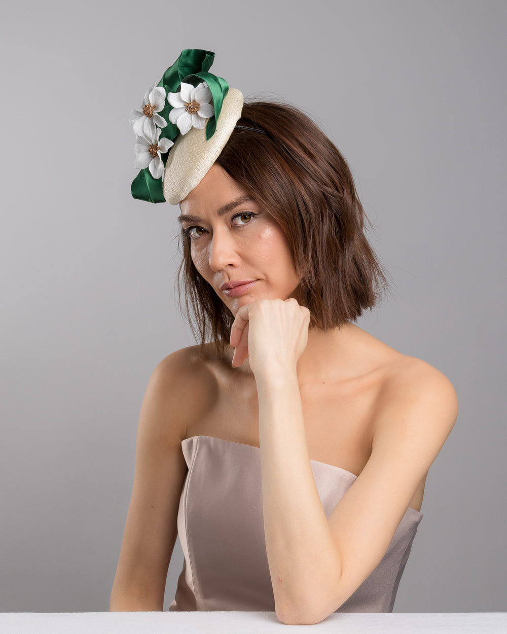 Female model is posing wearing Val, a button shaped, ivory sinamay fascinator worn to the front right side of the head. The fascinator is decorated with delicate white silk flowers and green sateen leaf swirls. Designed and made by Merve Bayindir millinery.