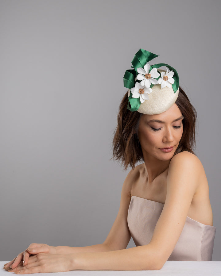 Female model is posing wearing Val, a button shaped, ivory sinamay fascinator worn to the front right side of the head. The fascinator is decorated with delicate white silk flowers and green sateen leaf swirls. Designed and made by Merve Bayindir millinery.