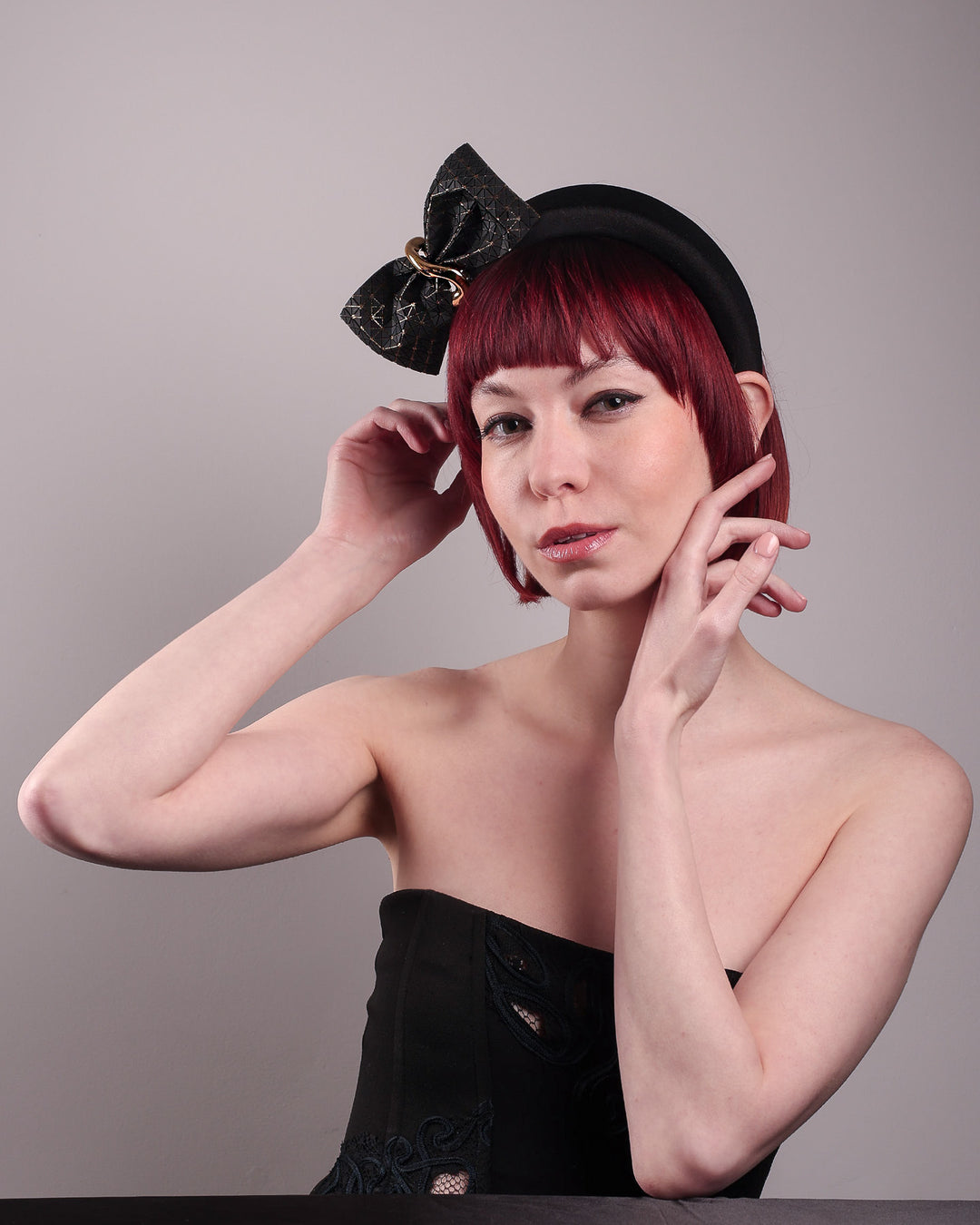 The model is wearing and showcasing the left front of the Valkyrie, which features a sculpted bow headband with a gold ring detail. This holds a structured bow in black faux leather with a gold metallic base. The bow is dramatically placed across a plush headband base cinched by the gold-tone ring. Designed by Merve Bayindir.
